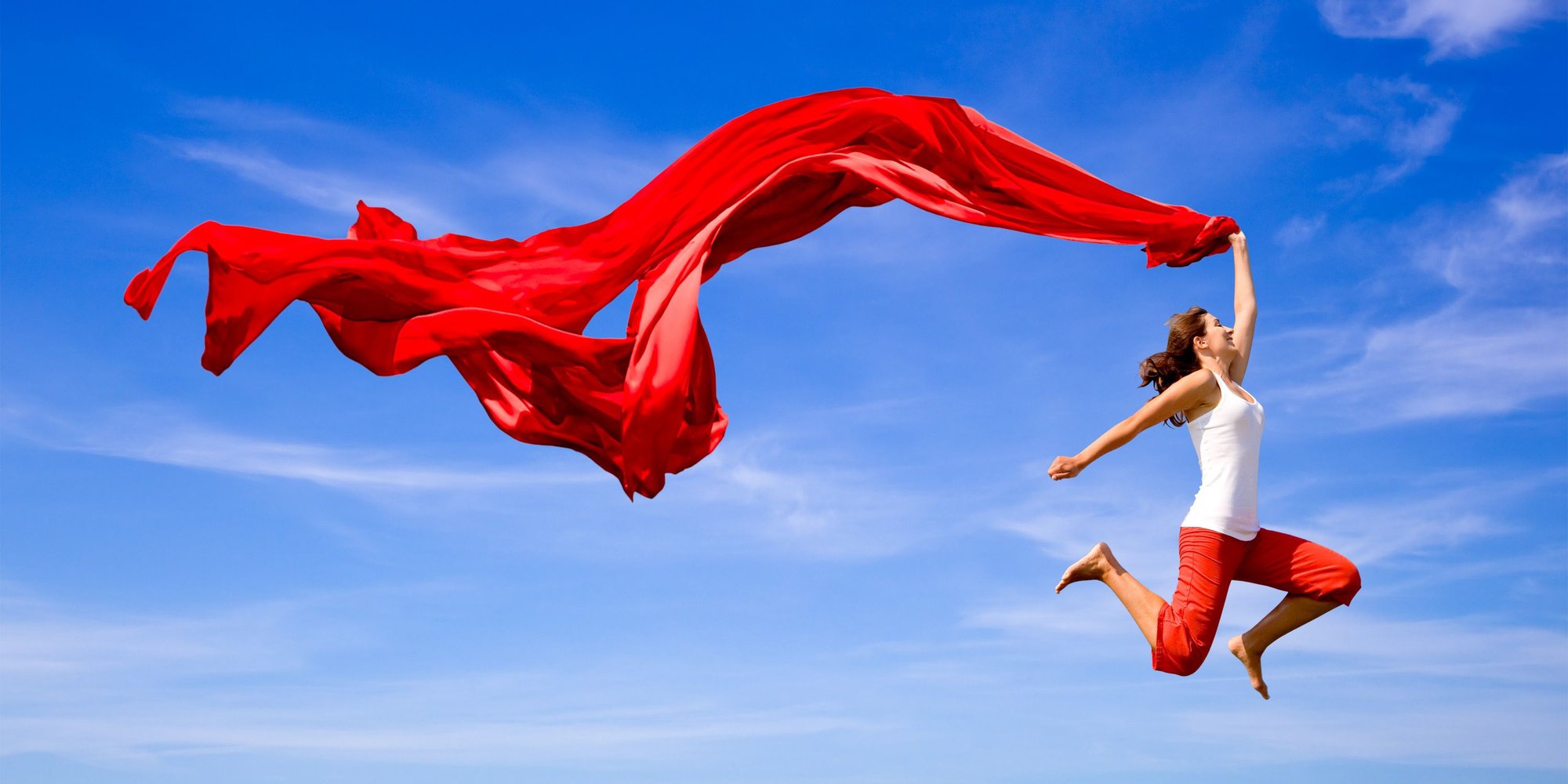 Kvinna hoppar med rött tygstycke mot blå himmel.