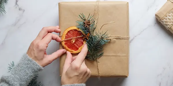Person slår in en julklapp, dekorerad med torkade apelsiner och en grankvist