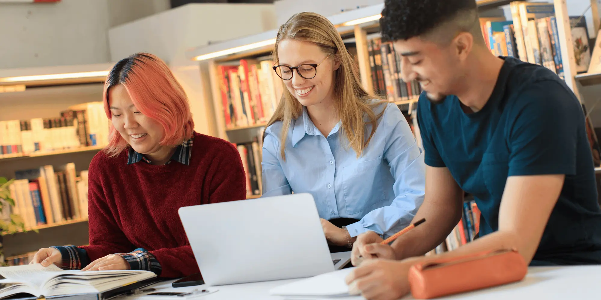 Tre studenter sitter vid ett bord i ett bibliotek och studerar tillsammans med böcker och en bärbar dator