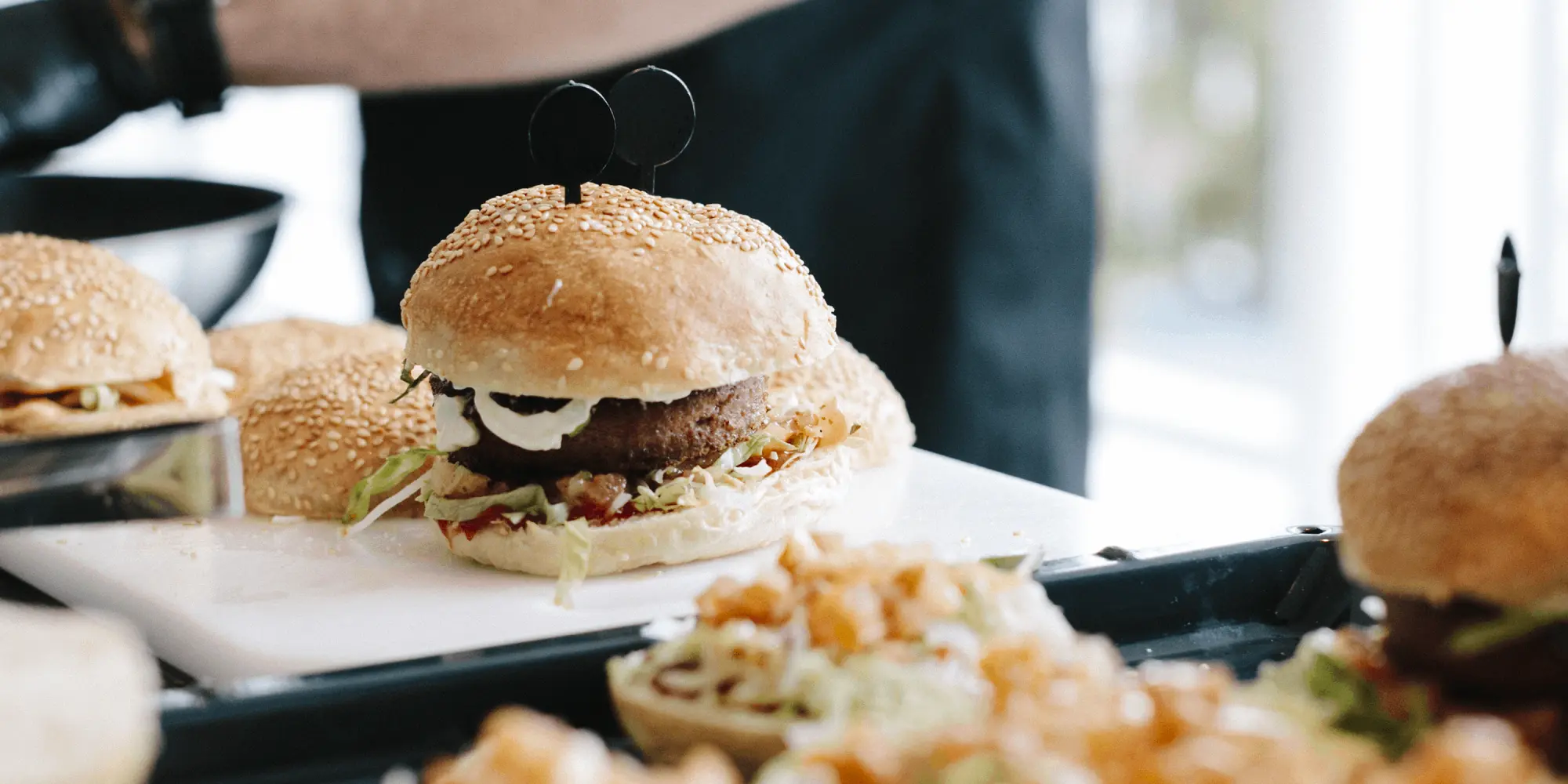 Hamburgare tillagas i ett restaurangkök. I förgrunden syns flera hamburgare med sesambröd, sallad och olika fyllningar. En person med svarta handskar arbetar vid en bänk i bakgrunden.