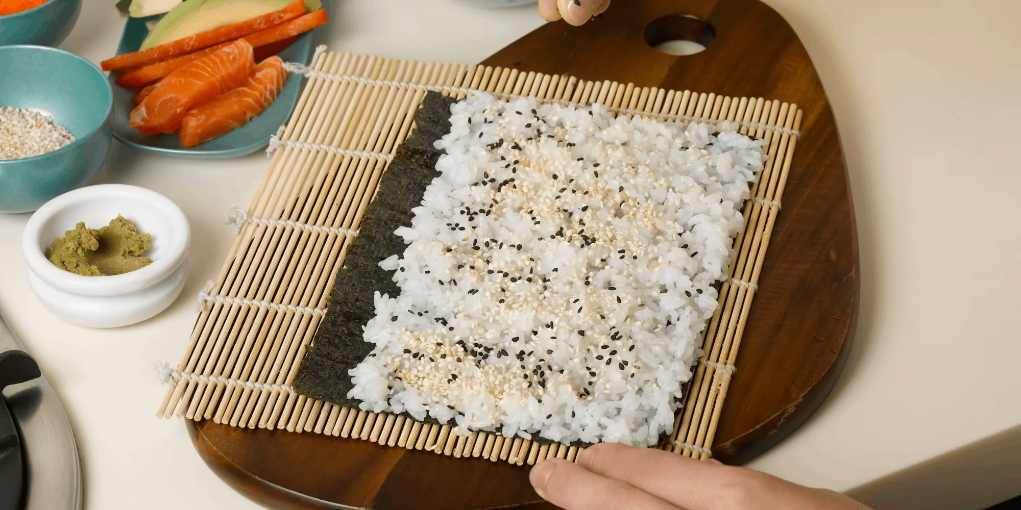 En person förbereder sushi genom att strö sesamfrön över ett lager ris som ligger på ett noriark på en bambumatta. Runtom finns skålar med ingredienser som lax, gurka, paprika, avokado, tobiko, wasabi och sesamfrön.