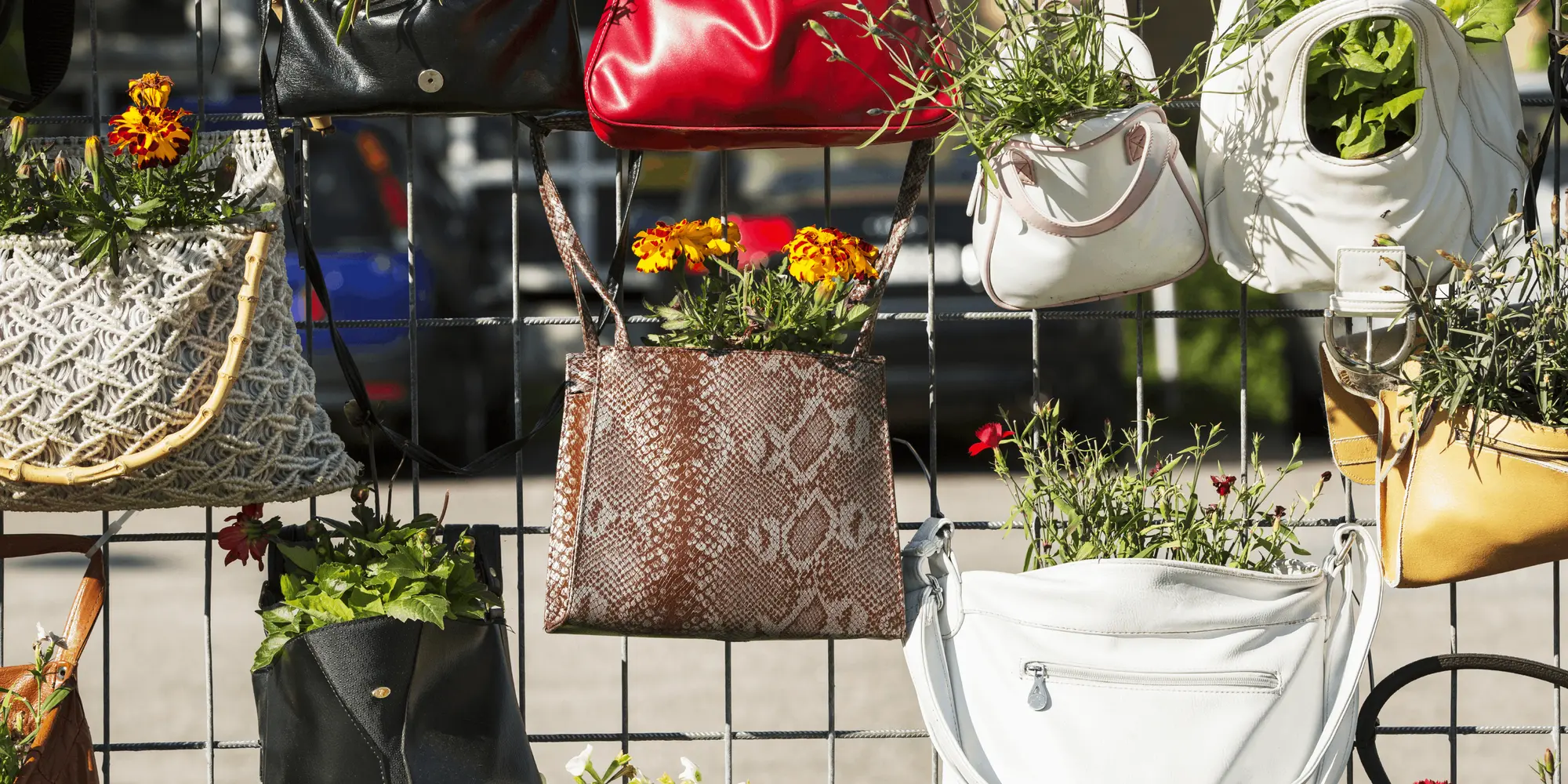 Återvunna handväskor som används som blomkrukor i en kreativ plantering mot ett stängsel. Färggranna växter spirar ur väskorna – ett exempel på återbruk och hållbar återvinning med estetisk och miljövänlig touch