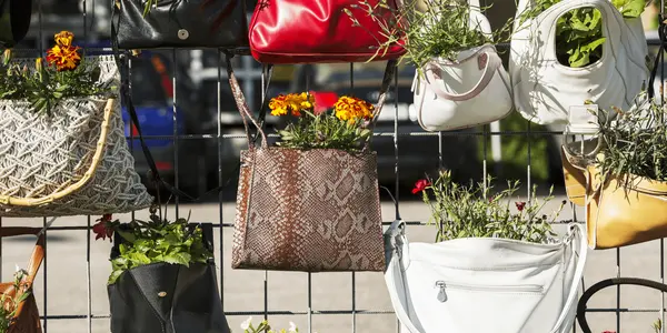 Återvunna handväskor som används som blomkrukor i en kreativ plantering mot ett stängsel. Färggranna växter spirar ur väskorna – ett exempel på återbruk och hållbar återvinning med estetisk och miljövänlig touch