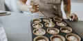 Kvinna i förkläde fyller muffinsformar med smet i en muffinsplåt med hjälp av en sked.