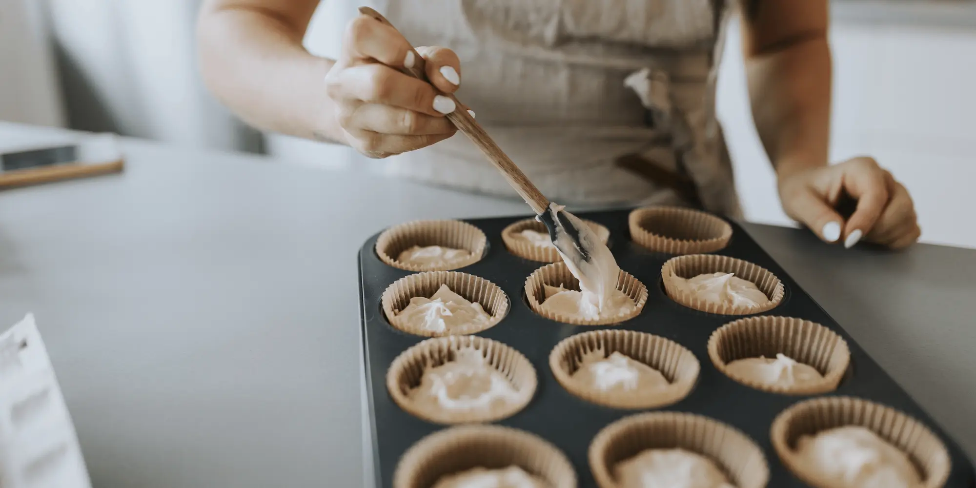 Kvinna i förkläde fyller muffinsformar med smet i en muffinsplåt med hjälp av en sked.
