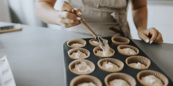 Kvinna i förkläde fyller muffinsformar med smet i en muffinsplåt med hjälp av en sked.