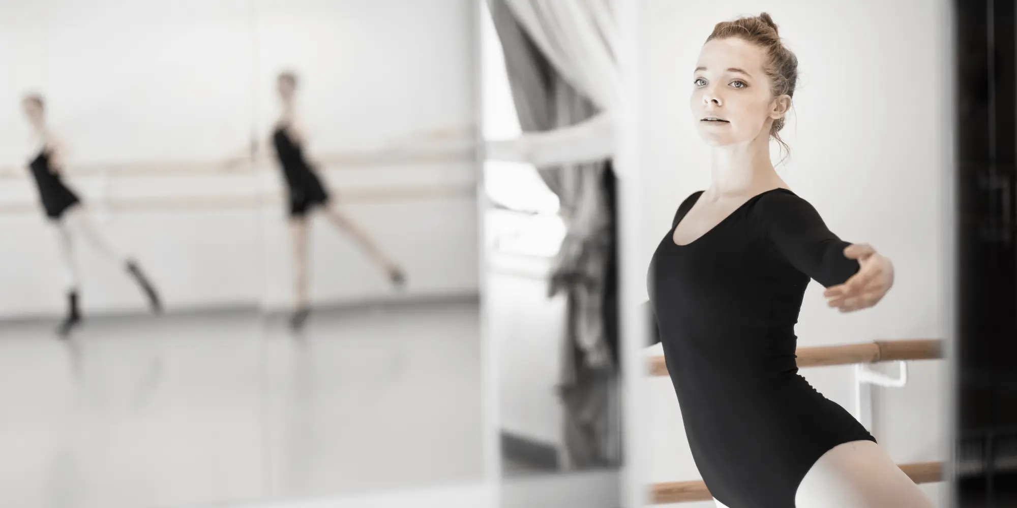 Ung kvinna i svart balettklädsel tränar vid balettstång i en ljus dansstudio.