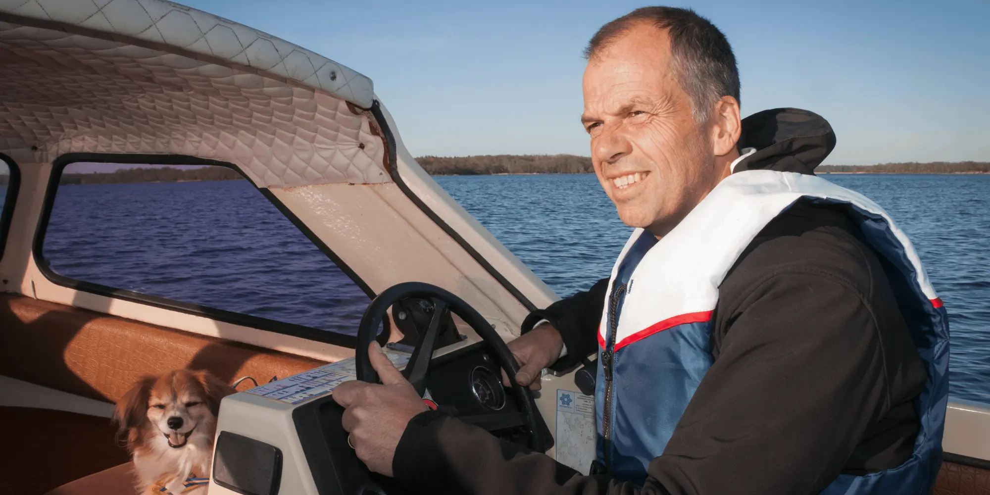 En man med flytväst styr en motorbåt i solsken medan en liten hund sitter avslappnat bredvid honom på en soffa inuti båten.