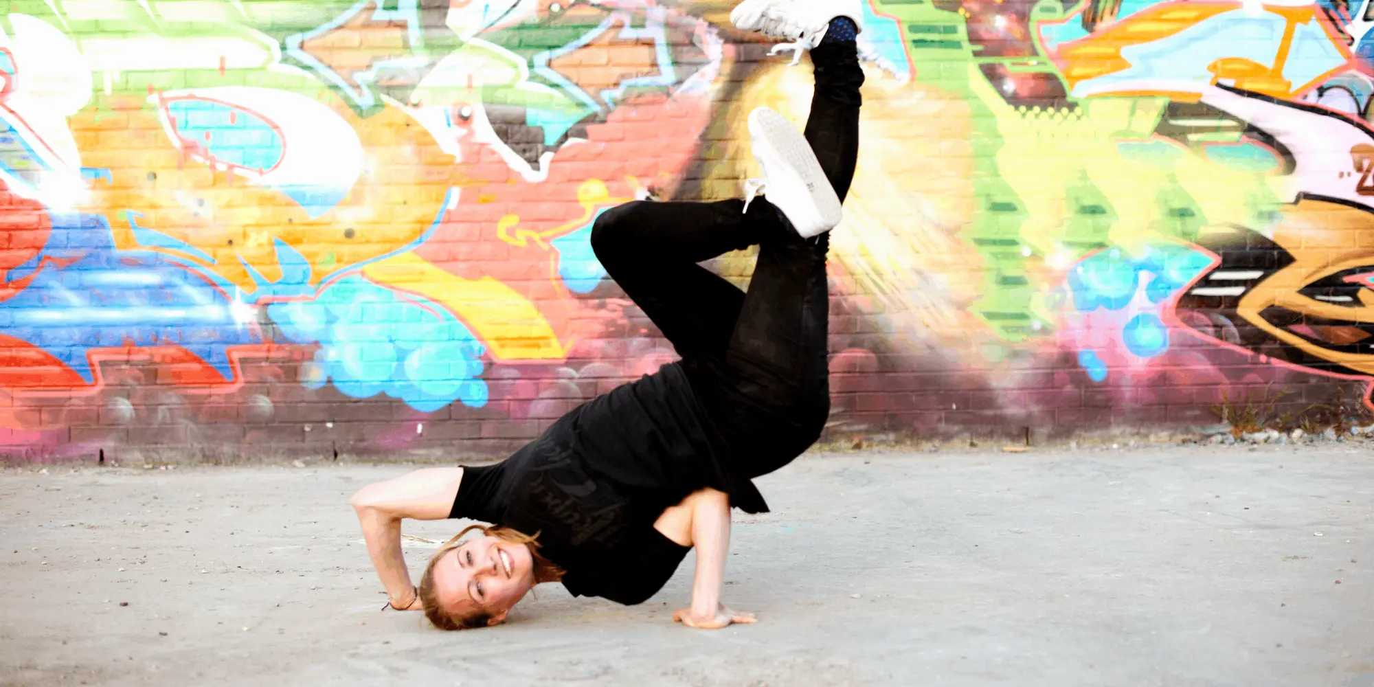 Ung person utför ett avancerat breakdance-move utomhus mot en färgglad graffitivägg