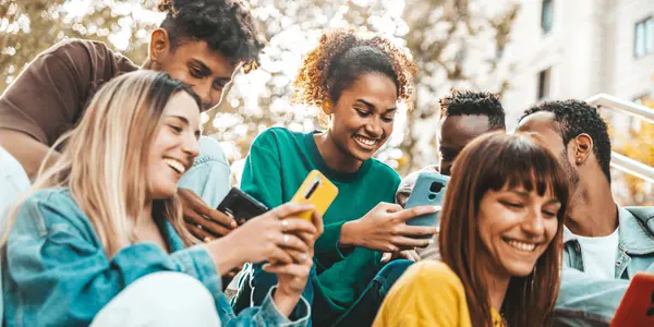 En grupp unga vuxna skrattar tillsammans medan de utforskar innehåll på sina mobiler i en utomhusmiljö.
