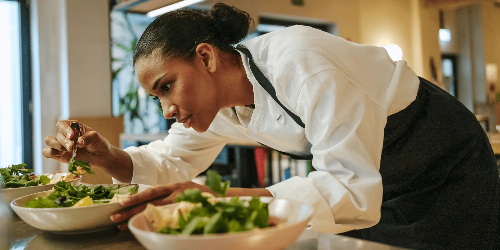 Kvinna i kockrock och förkläde fokuserar noggrant när hon dekorerar en salladsskål med tång. Framför henne står flera skålar fyllda med fräscha grönsaker och citronbitar i ett professionellt kök.