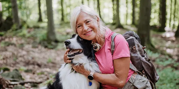 Äldre kvinna i vandringskläder sitter i en grönskande skog och kramar sin svartvita hund.