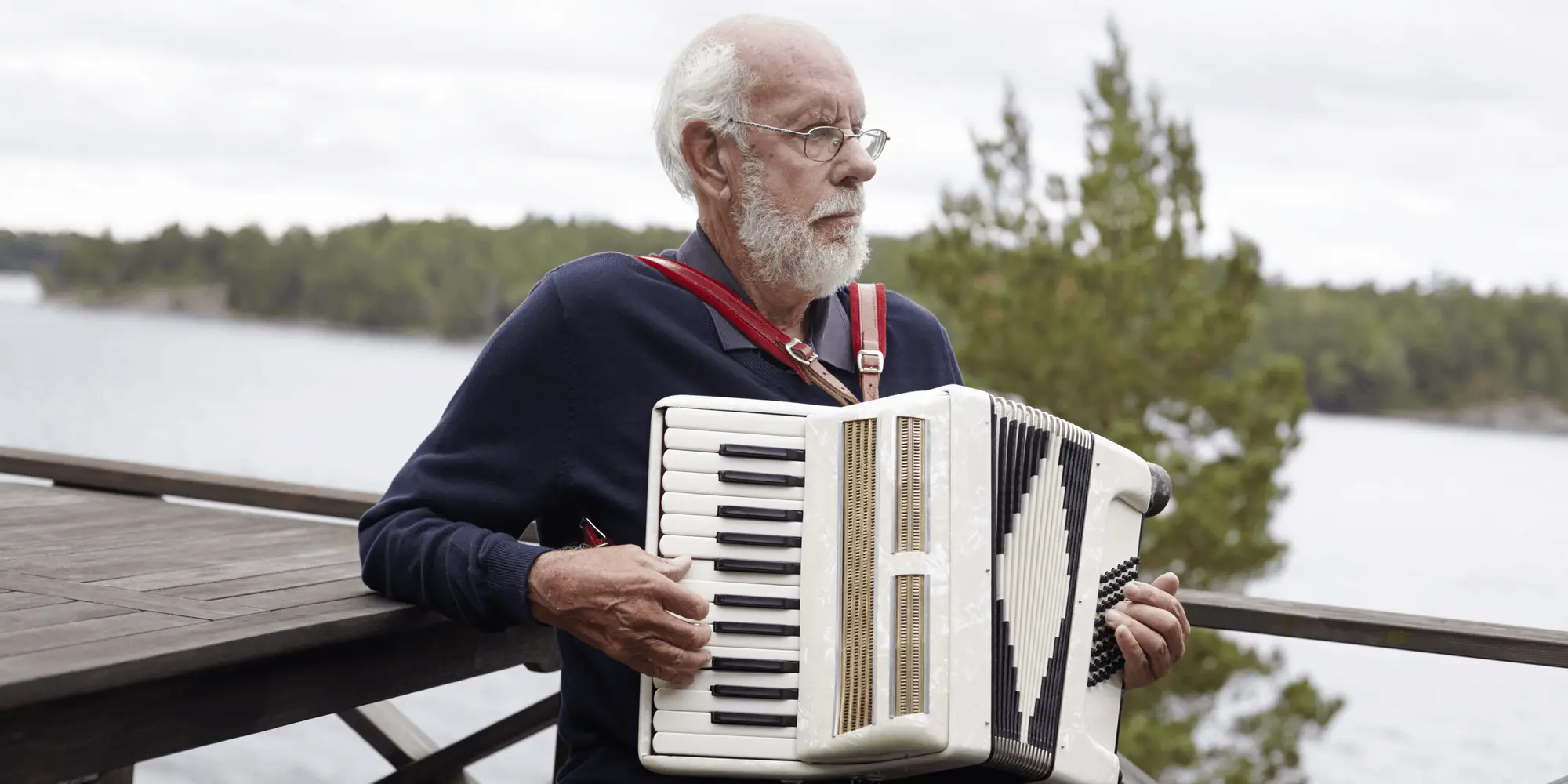Äldre man spelar dragspel vid vattnet med skog i bakgrunden