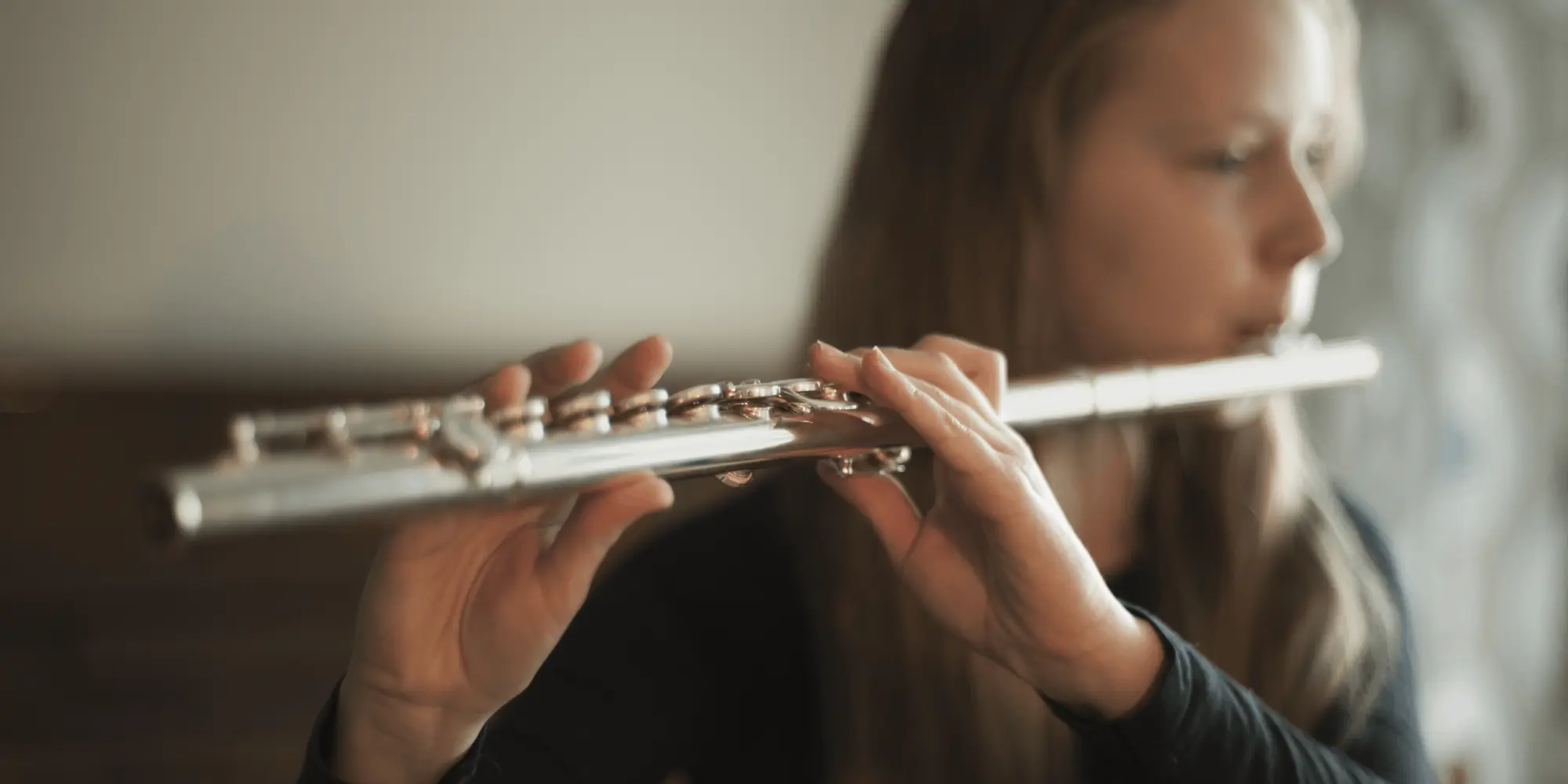 Kvinnlig flöjtist spelar tvärflöjt i ett mjukt upplyst rum, med fokus på händerna och instrumentet.