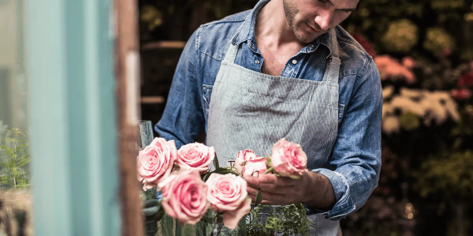 Florist i jeansskjorta och förkläde arrangerar rosa rosor i en blomsterbutik, omgiven av blommor i bakgrunden