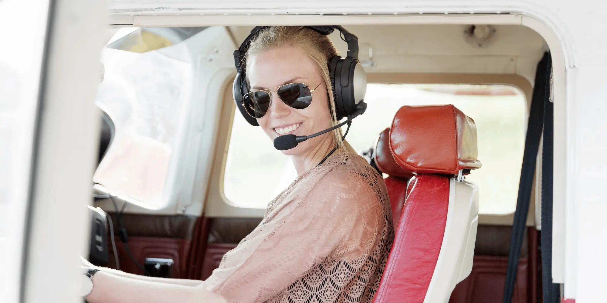 Leende kvinna med solglasögon och headset sitter i ett litet flygplan och håller i spakarna