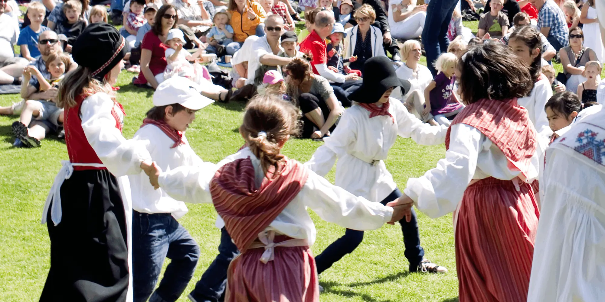 Barn i traditionella folkdräkter dansar ringdans utomhus på en gräsmatta inför en publik som sitter i solen