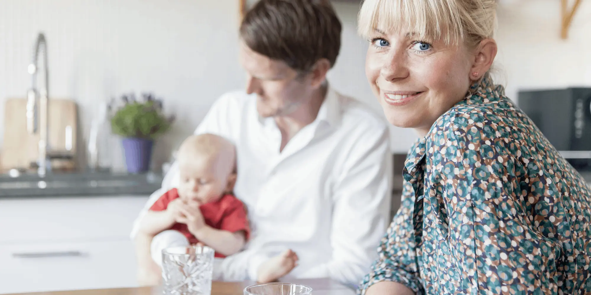Kvinna ler mot kameran vid ett köksbord, medan en man i bakgrunden håller en bebis i famnen. De befinner sig i ett ljust, modernt kök.
