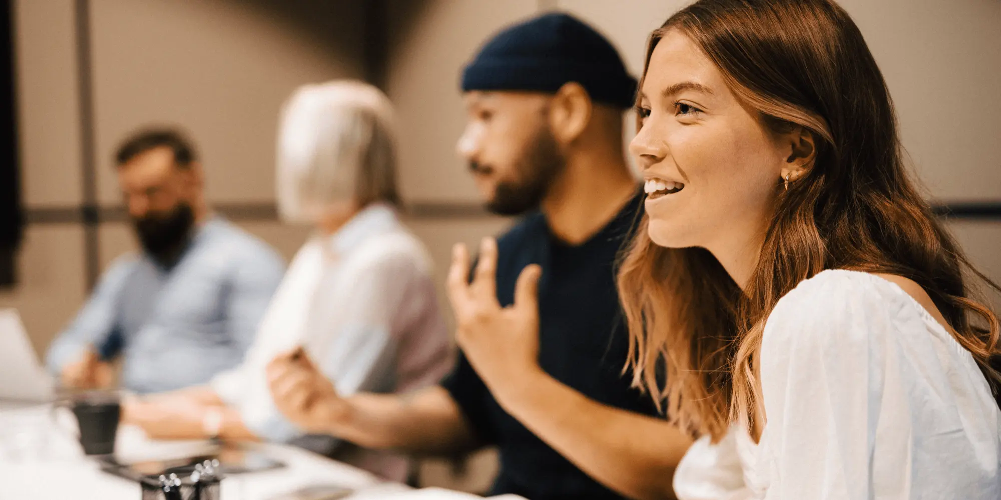 Fyra personer sitter vid ett konferensbord i ett modernt mötesrum. En kvinna i förgrunden ler och deltar aktivt i samtalet, medan övriga lyssnar och anteckna