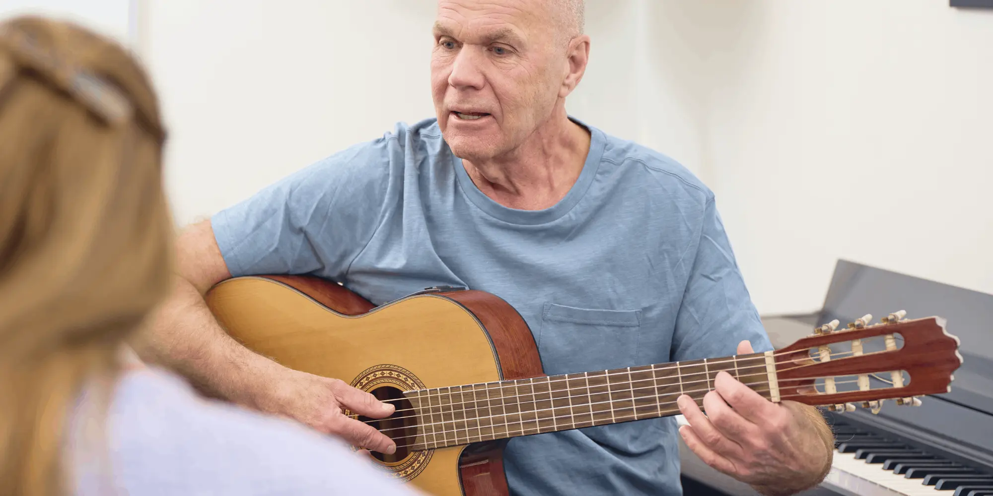 Äldre man undervisar i gitarrspel, sittandes med en akustisk gitarr i famnen och pratar med en elev i förgrunden.