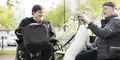Två personer sitter i en park, en i permobil och en på bänk som klappar en hund. Båda ser glada ut och samtalar i vårsolens sken, med grönskande träd i bakgrunden.