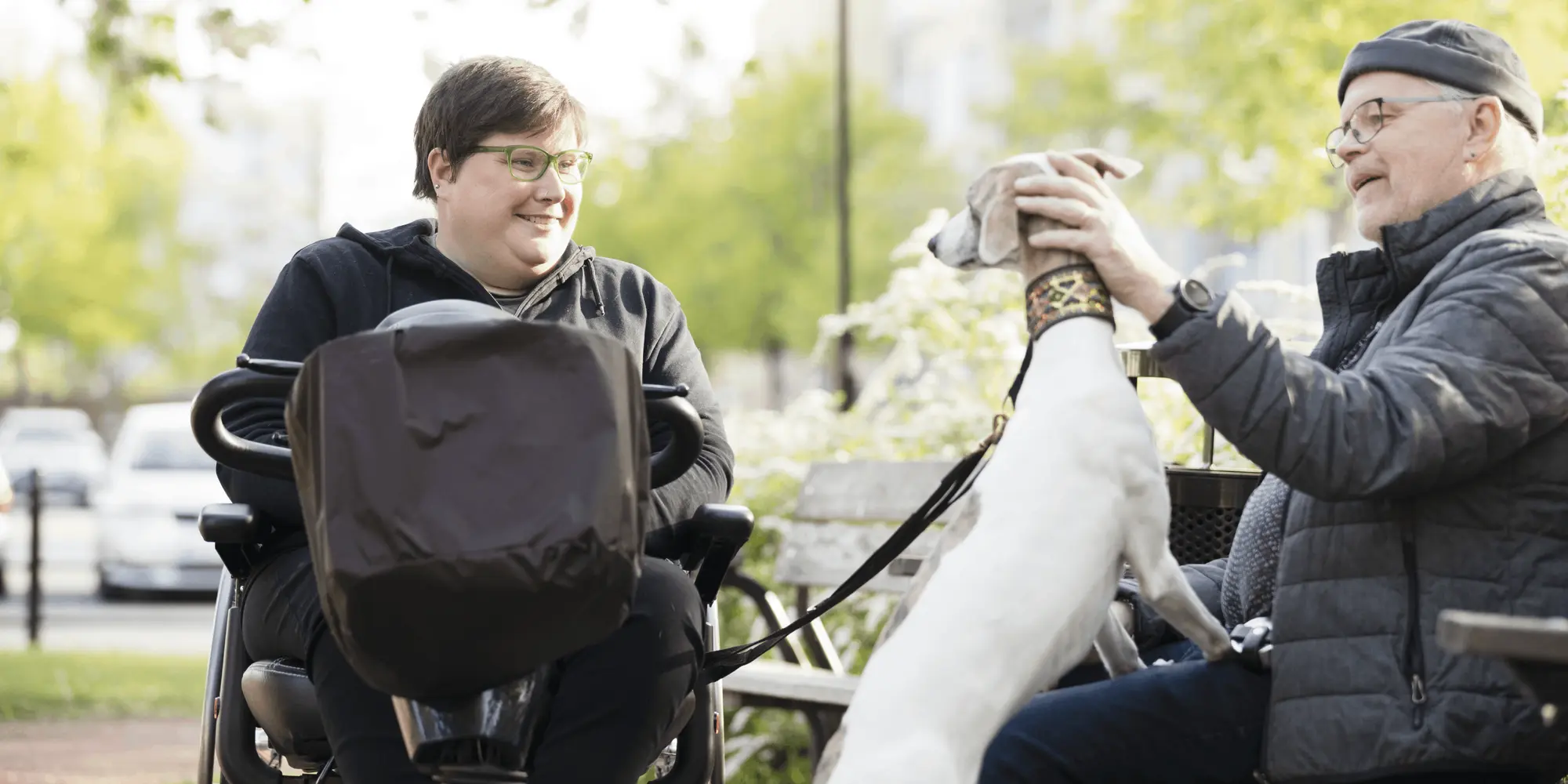 Två personer sitter i en park, en i permobil och en på bänk som klappar en hund. Båda ser glada ut och samtalar i vårsolens sken, med grönskande träd i bakgrunden.