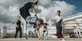 Ung man gör en akrobatisk breakdance-rörelse utomhus på ett tak, omgiven av tre vänner som hejar och ser imponerade ut