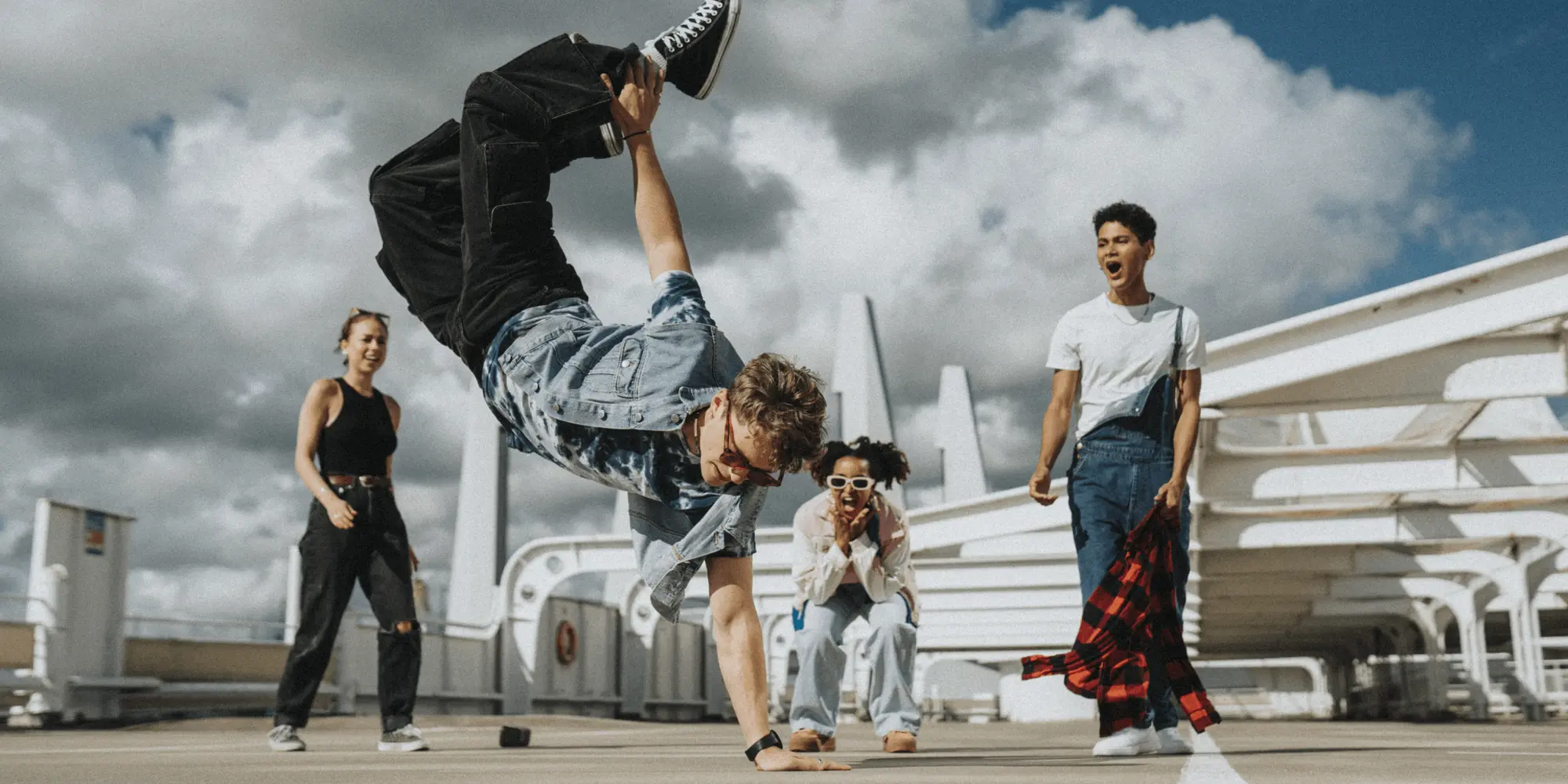 Ung man gör en akrobatisk breakdance-rörelse utomhus på ett tak, omgiven av tre vänner som hejar och ser imponerade ut
