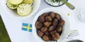 Svensk måltid med köttbullar, kokt potatis med gräslök och gräddfil i skålar på en vit duk. En liten svensk flagga pryder bordet, vilket förstärker känslan av traditionell svensk husmanskost.
