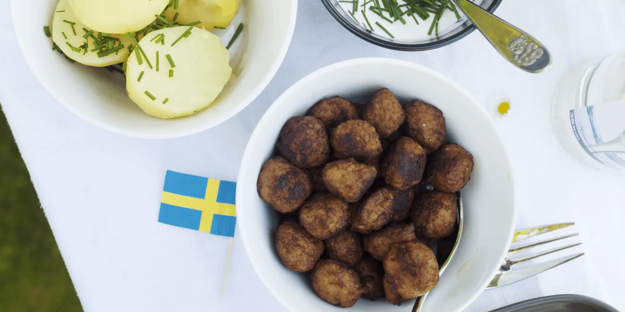 Svensk måltid med köttbullar, kokt potatis med gräslök och gräddfil i skålar på en vit duk. En liten svensk flagga pryder bordet, vilket förstärker känslan av traditionell svensk husmanskost.