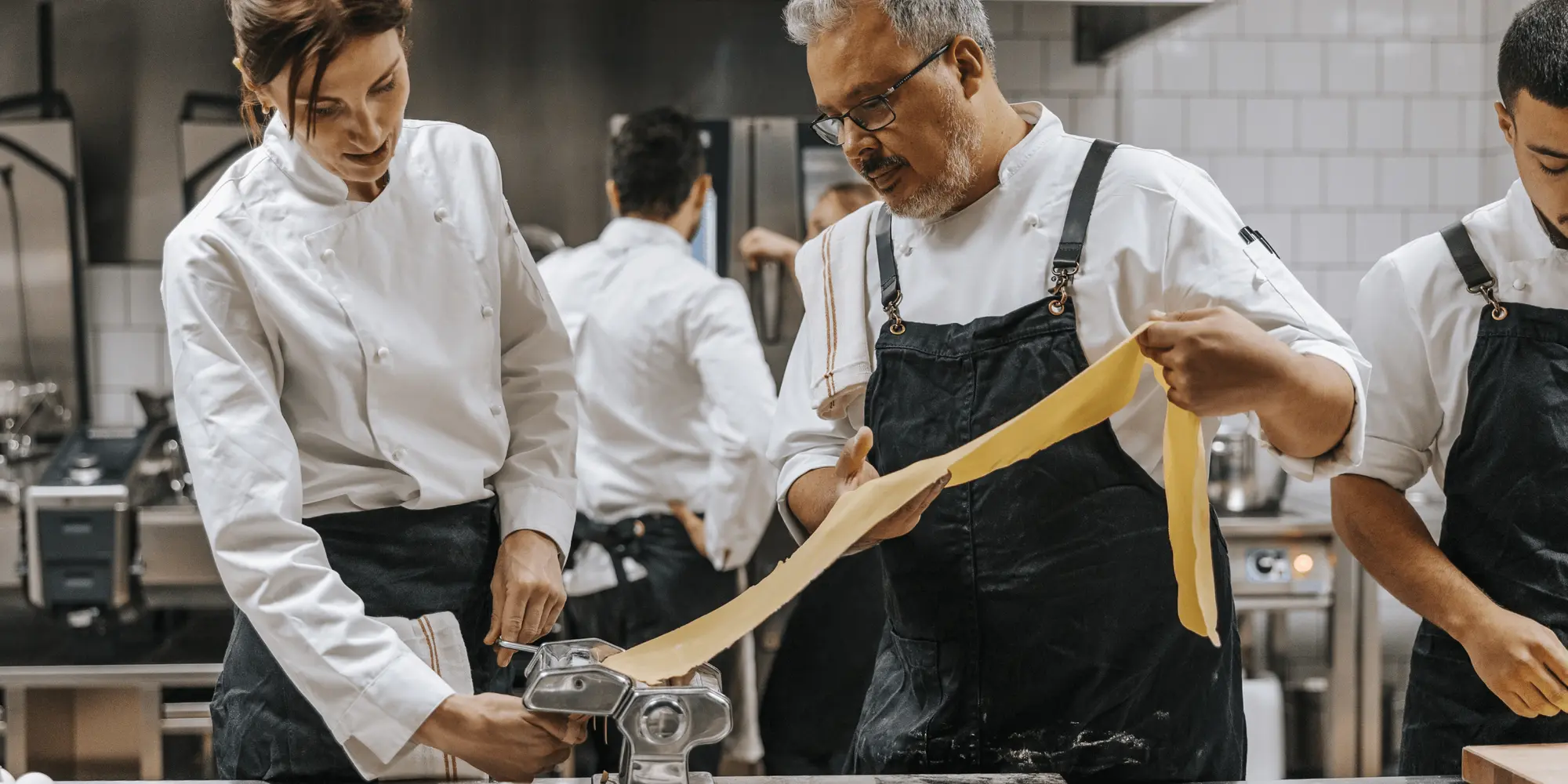 Två professionella kockar samarbetar i ett restaurangkök och vevar ut färsk pastadeg i en pastamaskin. Den ena håller i degen medan den andra matar den genom maskinen