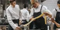 Två professionella kockar samarbetar i ett restaurangkök och vevar ut färsk pastadeg i en pastamaskin. Den ena håller i degen medan den andra matar den genom maskinen