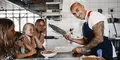En kock med tatuerade armar visar en knippe purjolök för en grupp nyfikna barn vid en köksbänk. Alla ser glada ut och befinner sig i ett professionellt kök.