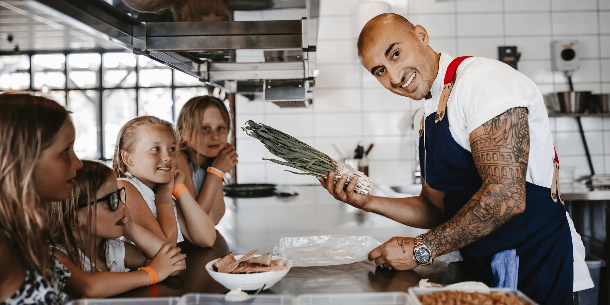 En kock med tatuerade armar visar en knippe purjolök för en grupp nyfikna barn vid en köksbänk. Alla ser glada ut och befinner sig i ett professionellt kök.