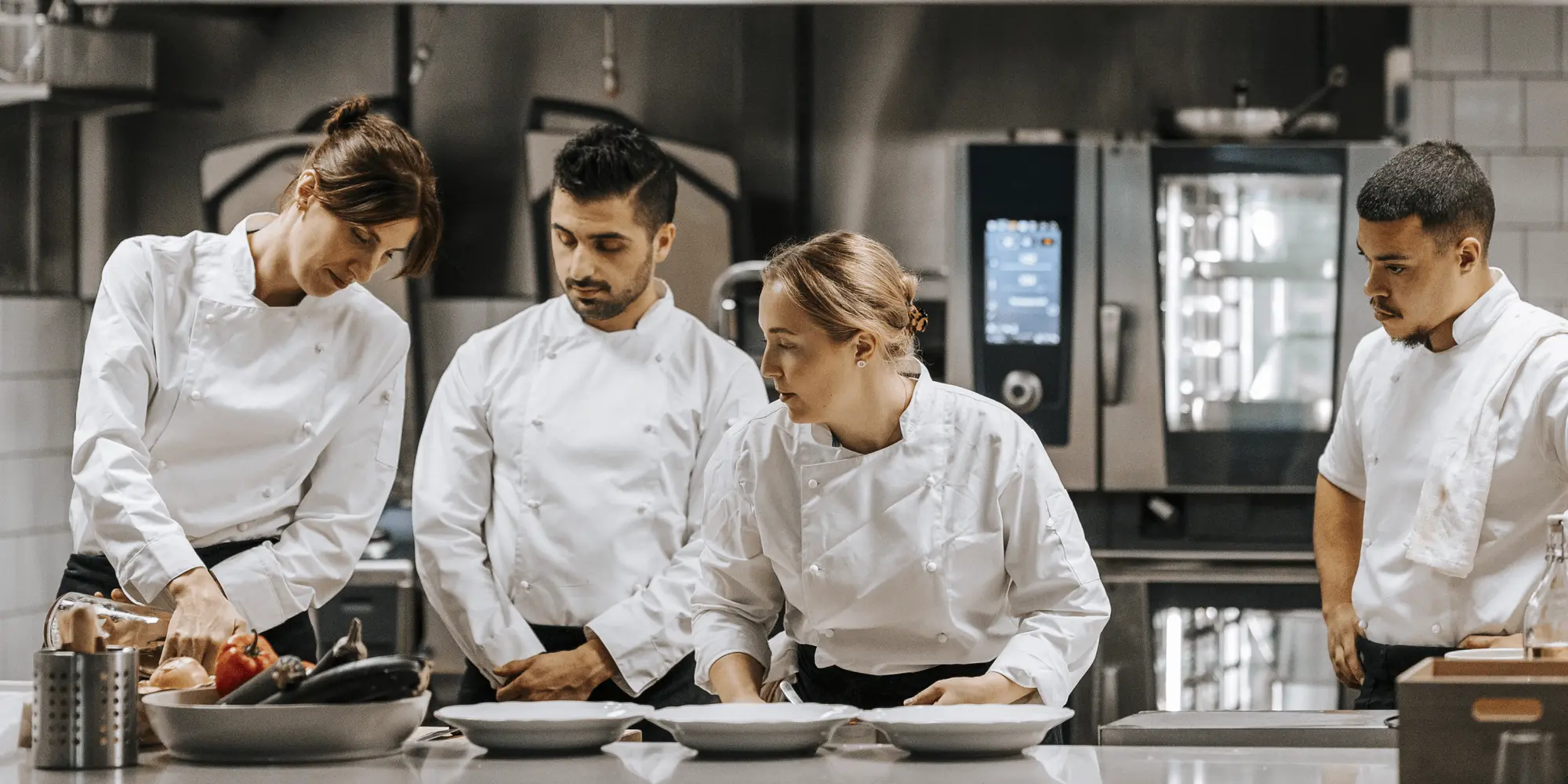 Fyra personer i kockkläder står tillsammans i ett restaurangkök och diskuterar matlagning. En kvinna visar något på ett skärbräde med grönsaker.