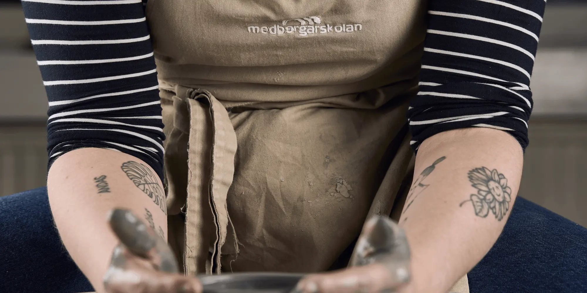En person med tatueringar på armarna arbetar med lera på en drejskiva. Personen bär ett förkläde med texten "Medborgarskolan" och en randig tröja. Fokus ligger på händerna som formar leran.