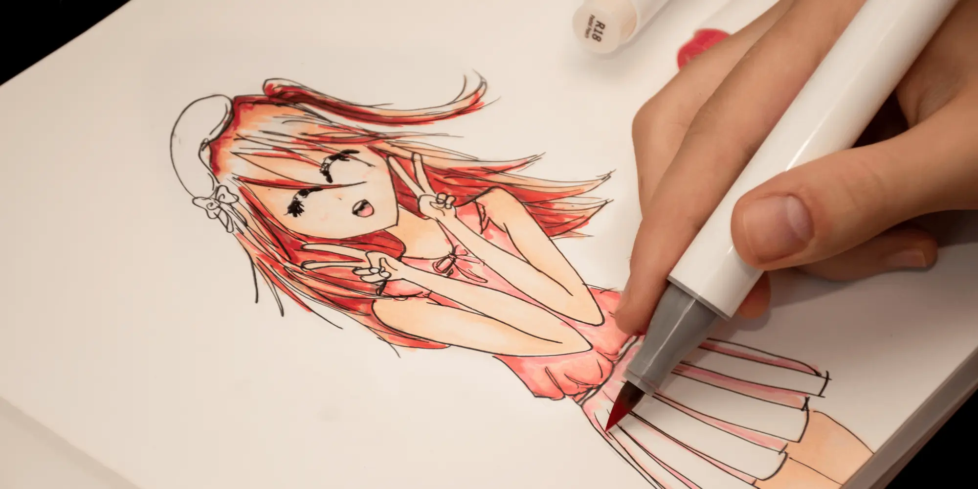 En person färglägger en teckning i manga-stil med en markerpenna. Motivet föreställer en flicka med långt rött hår, rosa topp och kjol som gör ett V-tecken med båda händerna.