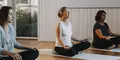 Tre kvinnor sitter i meditationsställning på yogamattor i ett ljust rum med trägolv och stora fönster. De har stängda ögon och ser fokuserade ut, med händerna vilande på knäna. Atmosfären är lugn och avslappnad.
