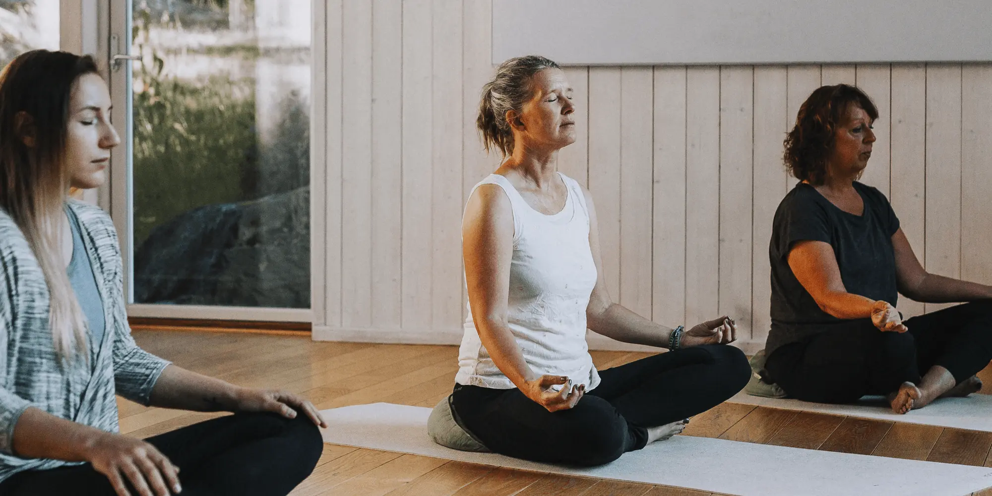 Tre kvinnor sitter i meditationsställning på yogamattor i ett ljust rum med trägolv och stora fönster. De har stängda ögon och ser fokuserade ut, med händerna vilande på knäna. Atmosfären är lugn och avslappnad.