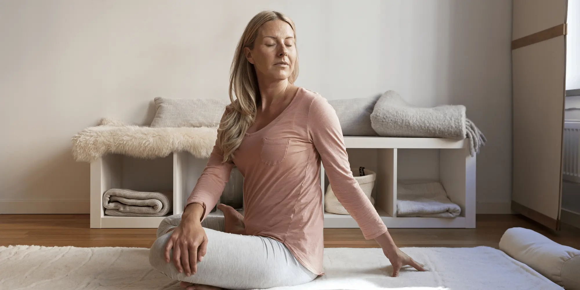 En kvinna sitter på en yogamatta i ett lugnt rum, med ögonen stängda och kroppen vriden i en sittande twistposition. Hon bär en ljusrosa tröja och ljusgrå byxor. Bakom henne syns en hylla med hoprullade filtar och korgar, vilket bidrar till en harmonisk o