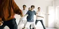 En grupp unga personer dansar tillsammans i ett ljust rum, leende och med energiska rörelser, ledda av en person i röd tröja i förgrunden