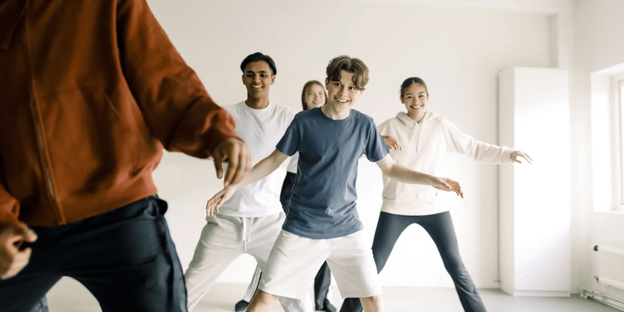 En grupp unga personer dansar tillsammans i ett ljust rum, leende och med energiska rörelser, ledda av en person i röd tröja i förgrunden