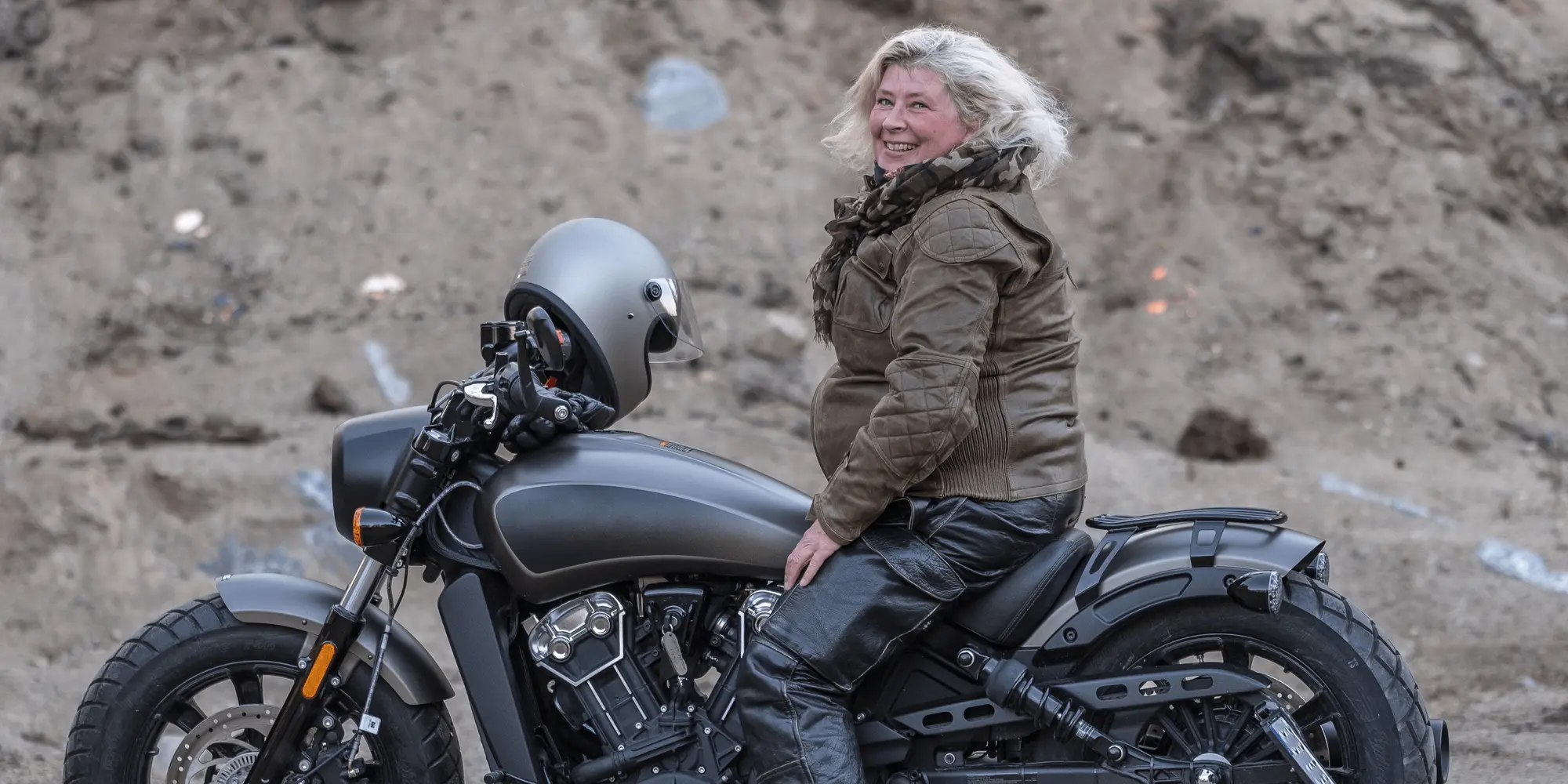 Leende kvinna i motorcykelutrustning sitter på en svart motorcykel mot en bakgrund av grus och sten