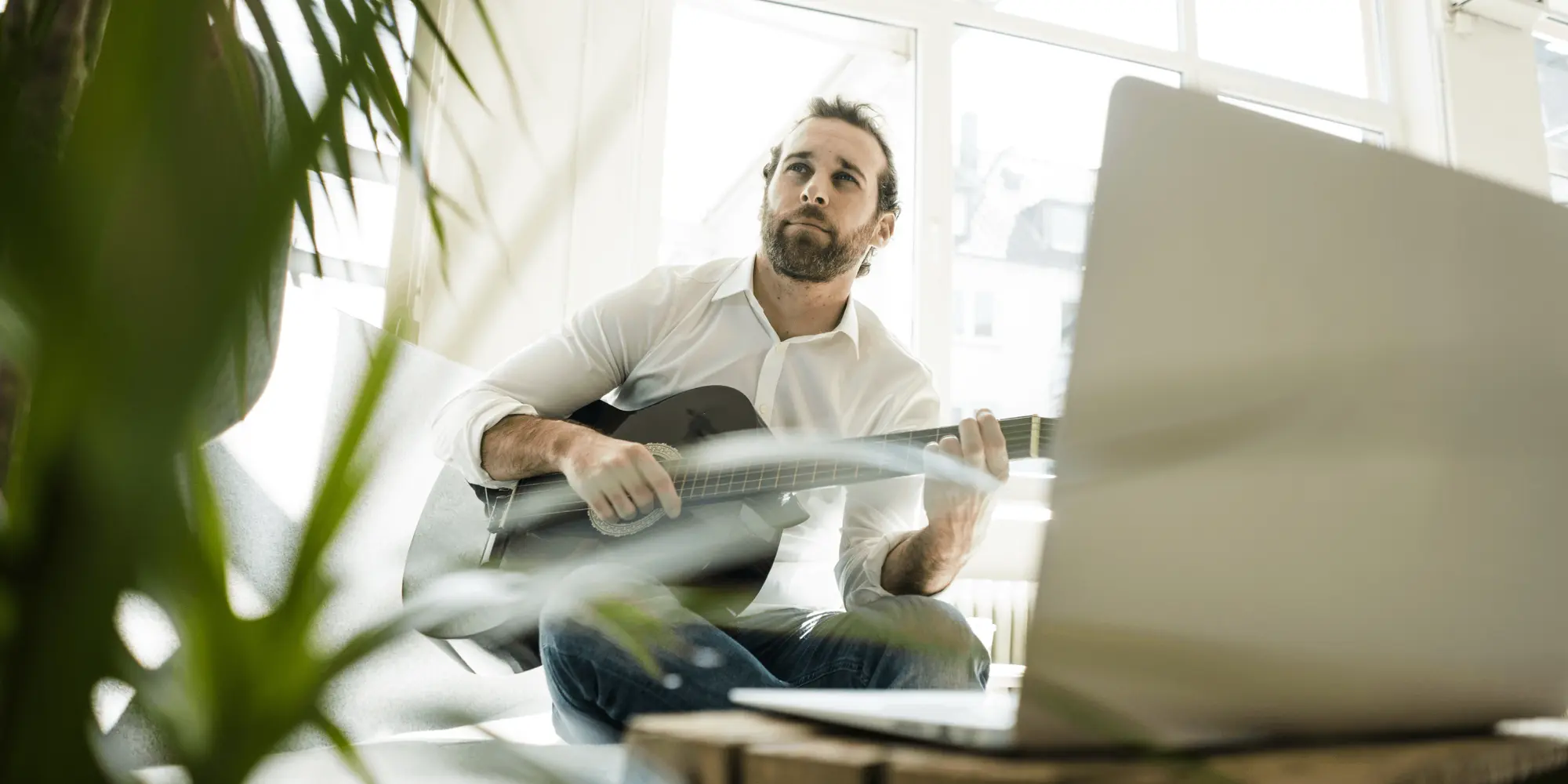 Man sitter i ett ljust rum och spelar gitarr framför en laptop