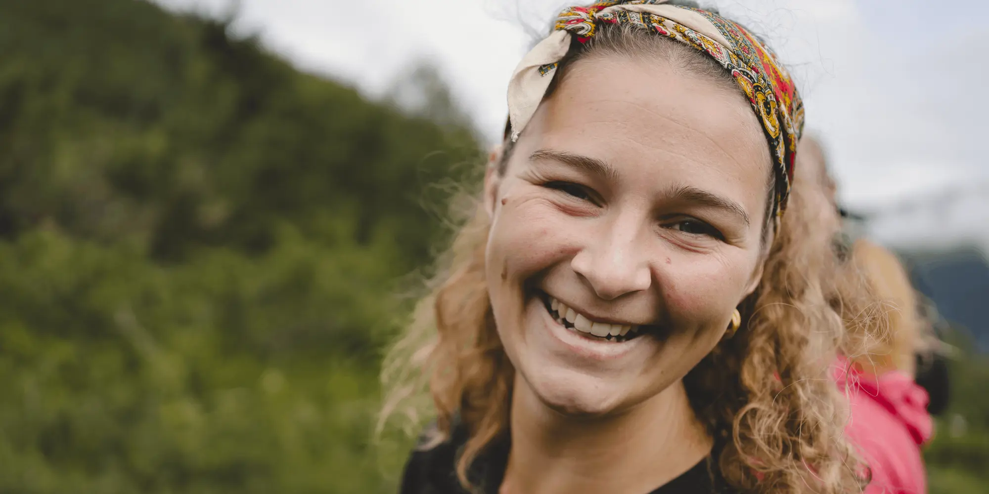 Närbild på en leende kvinna med lockigt hår och färgglad sjal i håret, stående utomhus med grönskande natur i bakgrunden