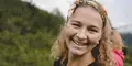 Närbild på en leende kvinna med lockigt hår och färgglad sjal i håret, stående utomhus med grönskande natur i bakgrunden