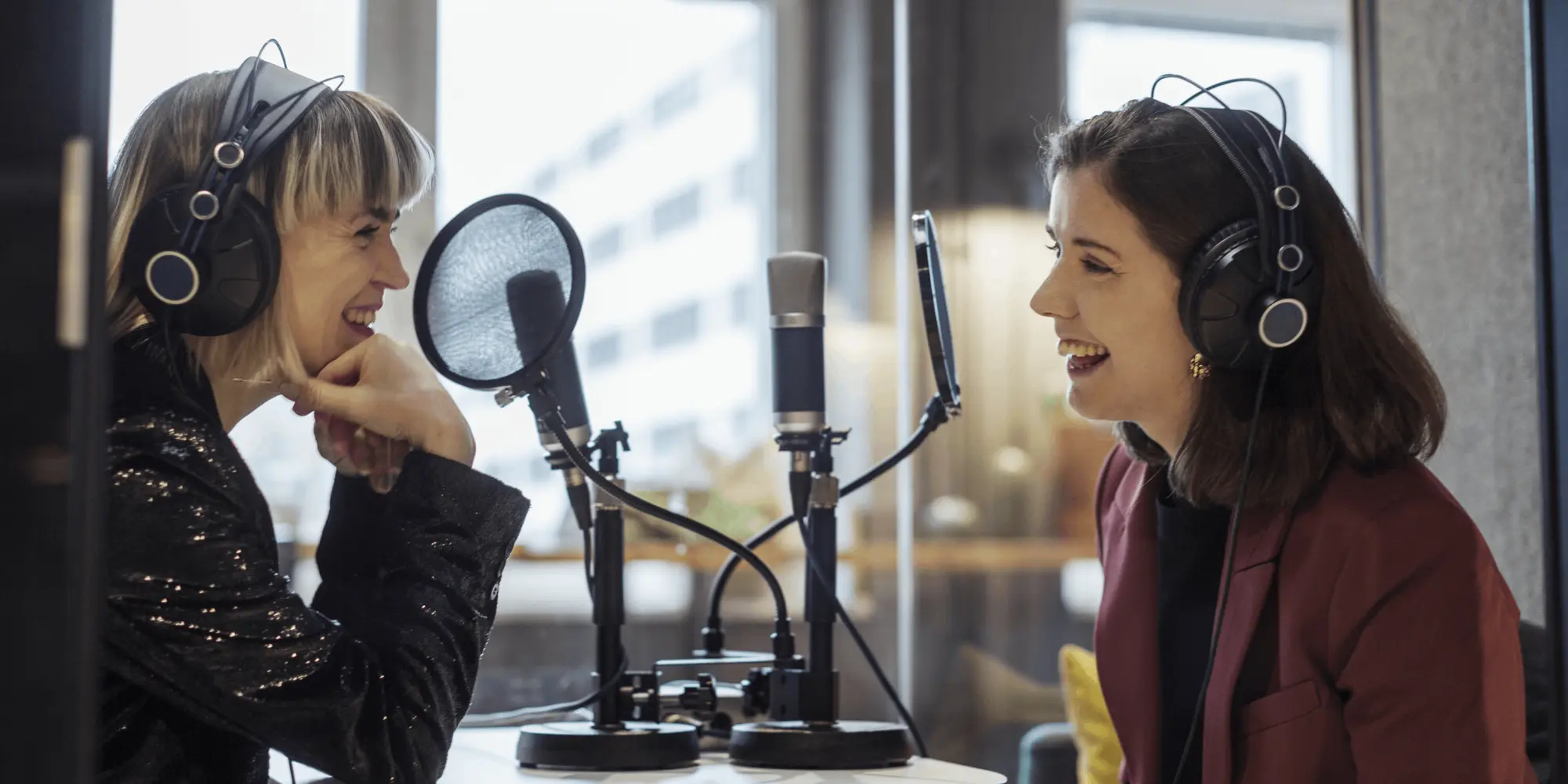 Två kvinnor med hörlurar sitter i en studio och spelar in ett samtal framför mikrofoner – poddinspelning eller radioproduktion i en kreativ miljö.