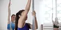 Kvinnor tränar pole dance i en ljus träningsstudio. De håller i varsin stång och fokuserar på övningen