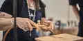 Närbild på en person som använder en gasbrännare för att arbeta med metall på en värmetålig stenplatta. Personen bär ett mörkblått förkläde, flera ringar och ett halsband