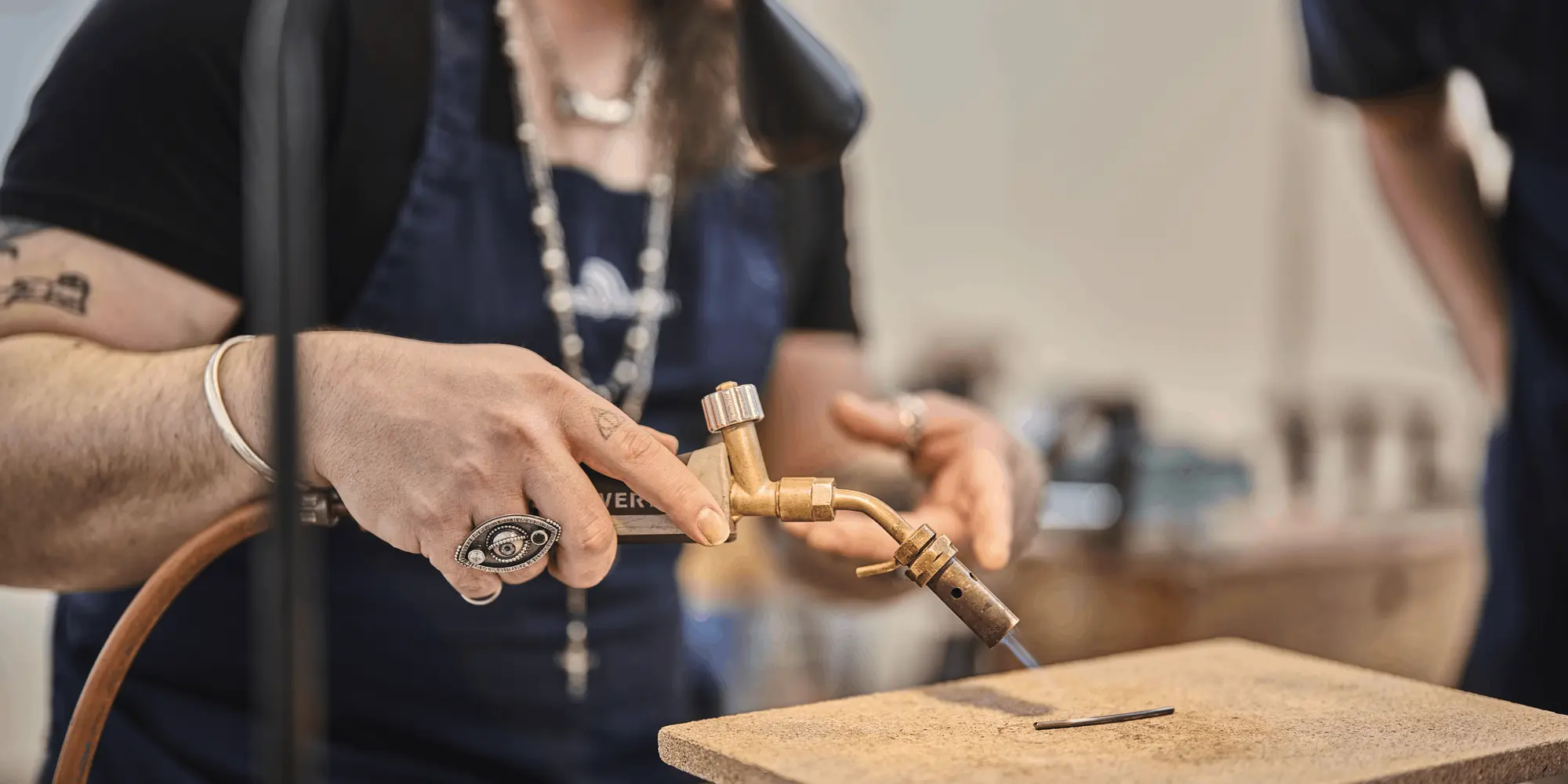 Närbild på en person som använder en gasbrännare för att arbeta med metall på en värmetålig stenplatta. Personen bär ett mörkblått förkläde, flera ringar och ett halsband